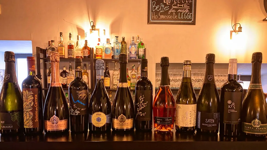 La mensola dietro il banco: bottiglie di Franciacorta, vermentino e cannonau in fila, e dietro lo scaffale dei distillati.