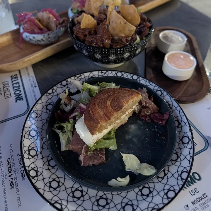 Brioche sfogliata farcita con carne salata e insalata, servita in un piatto scuro con le salse a fianco.