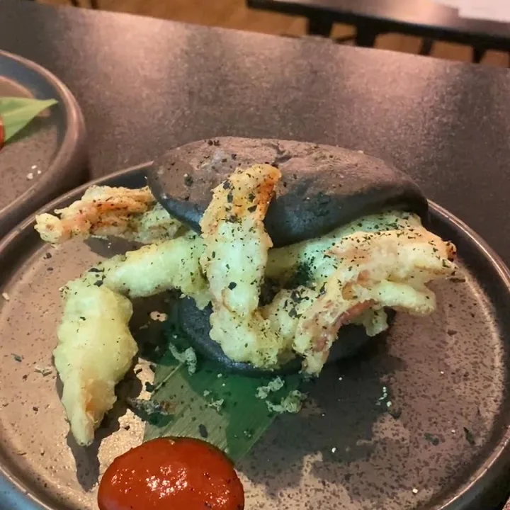 Gamberi in tempura appoggiati su un panino nero al carbone, con una salsa rossa nel piatto di ceramica.