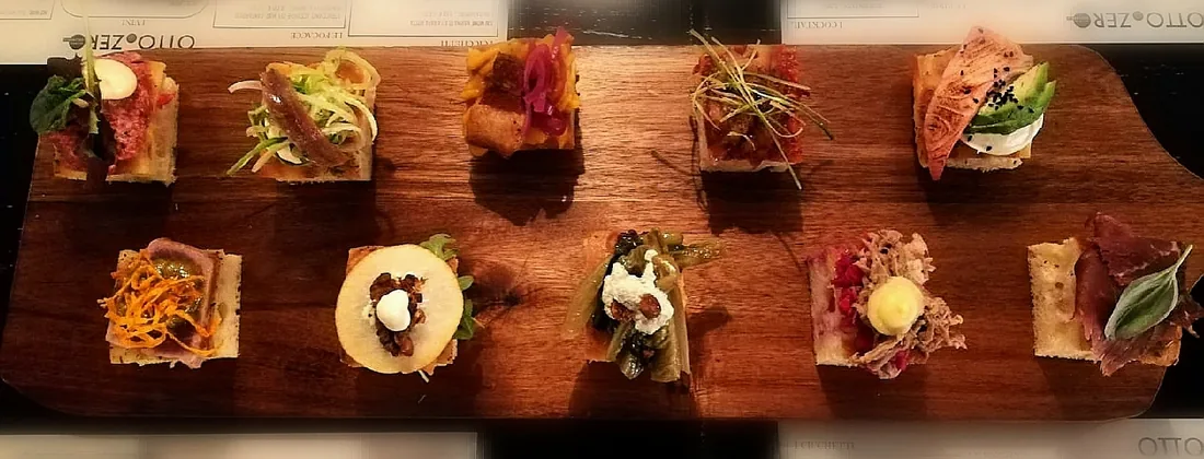 Un tagliere di legno lungo con dieci cicchetti allineati su due file: salmone e avocado, tartare, salumi, verdure, formaggi.